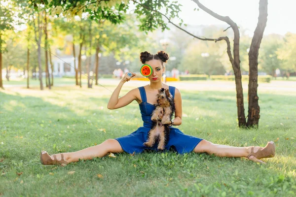 Bright makeup beautiful girl with Yorkshire Terrier seating on the grass holding watermelon lollipop.