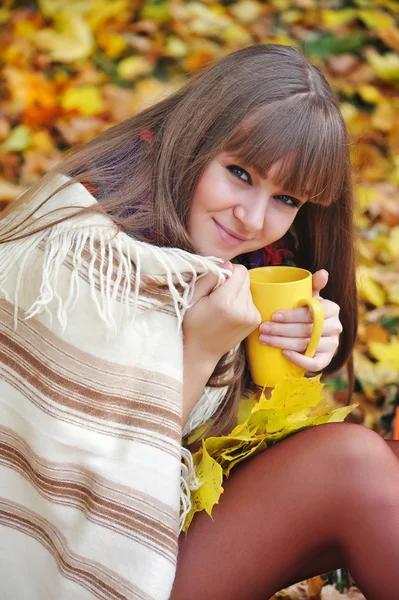 Young beautiful woman that holds the cup of tea in hands on a background autumn leaves