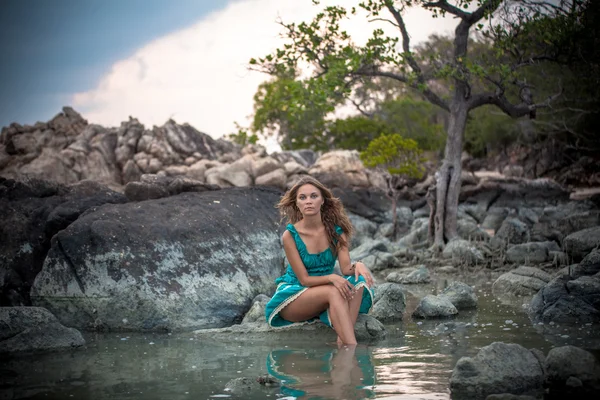 Young beautiful woman in long turquoise dress sitting on a stone by the sea