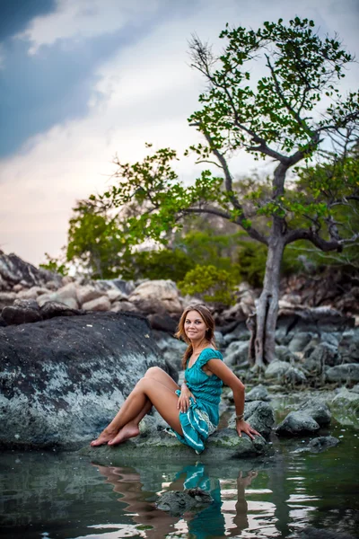 Young beautiful woman in long turquoise dress sitting on a stone by the sea