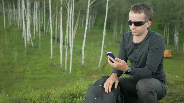 Young man in sunglasses pulls the phone out of the bag and talking. Against the background  beautiful nature: green trees  grass