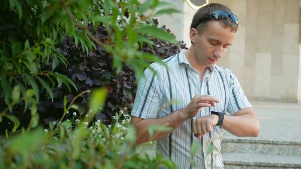 Outdoor portrait of modern young man with smart watch.  in sunglasses and jacket. Around a lot  green plants