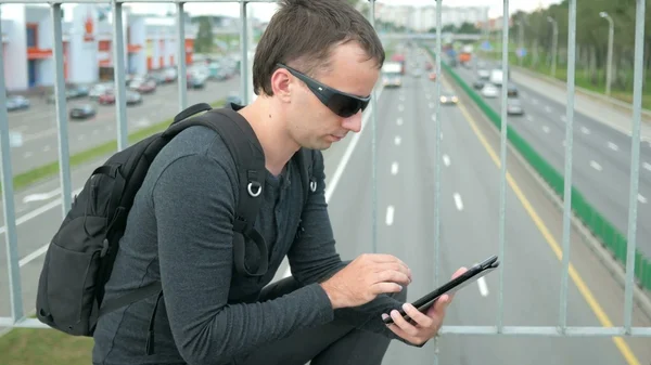 Outdoor portrait of modern young man with digital tablet in the street. The in glasses backpack sitting on the bridge. Bottom drive cars