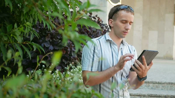 Outdoor portrait of modern young man with digital tablet.  in sunglasses and jacket. Around a lot  green plants