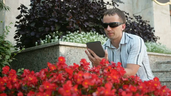 Outdoor portrait of modern young man with digital tablet.  in sunglasses and jacket. Near a lot  red flowers