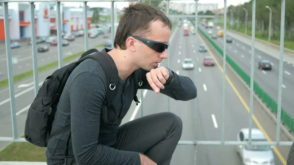 Outdoor portrait of modern young man with smart watch in the street. The  in glasses  backpack sitting on the bridge. Bottom drive cars