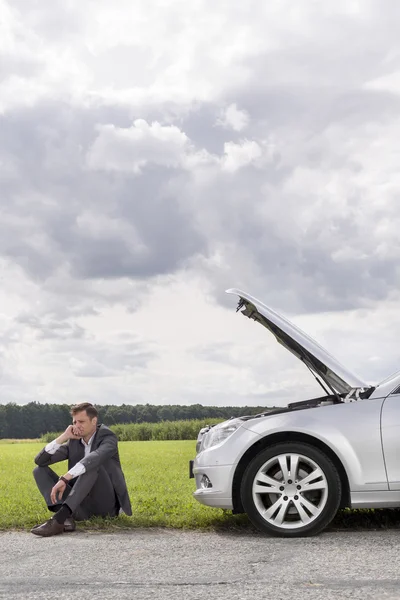Young businessman sitting by broken down car