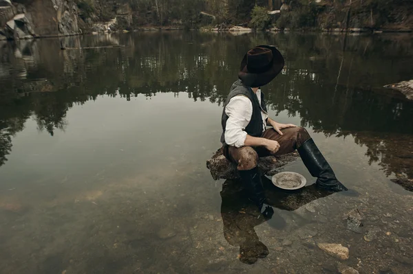 Unshaved golddigger washes gold in the lake with rocky bank