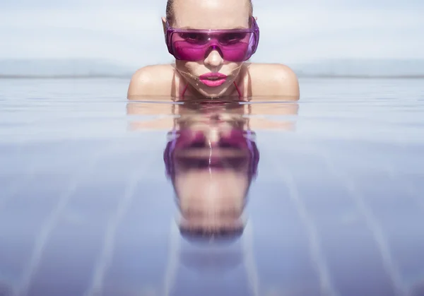 Face closeup of sexy young woman wearing purple sunglasses looking at camera in infinity rooftop swimming pool on a sunny day over blue sky and green trees landscape