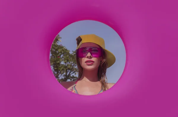 Pretty young woman wearing bikini, yellow hat and purple sunglasses with pink inflatable ring posing in infinity rooftop pool on a sunny day over blue sky and green trees landscape