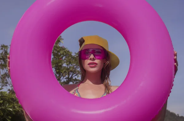 Pretty young woman wearing bikini, yellow hat and purple sunglasses with pink inflatable ring posing in infinity rooftop pool on a sunny day over blue sky and green trees landscape