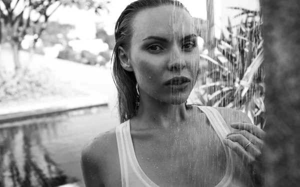 Front view of sexy blonde woman in bikini and white see-through tank top taking outdoors shower on the rooftop surrounded with tropical garden - black and white photo