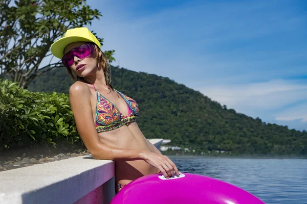 Pretty young woman wearing bikini, yellow hat and purple sunglasses with pink inflatable ring posing in infinity rooftop pool on a sunny day over blue sky and green trees landscape