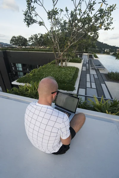 Top view of young man back holding laptop computer on his knees while sitting on a rooftop over infinity swimming pool and sky background. Freelance and technology