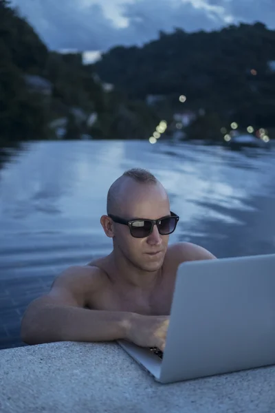 Outdoors portrait of handsome young man in sunglasses with laptop computer while standing in a rooftop infinitiy swimming pool over blurred green mountain and sky background during evening. Freelance and technology