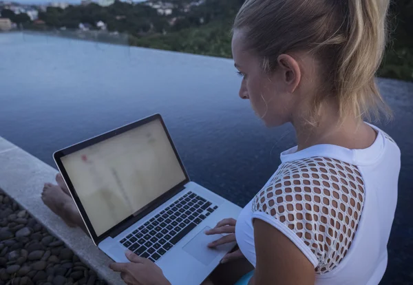 Side view of pretty blonde woman in white top with laptop computer on her knees sitting on a rooftop infinity swimming pool. Freelance and technology