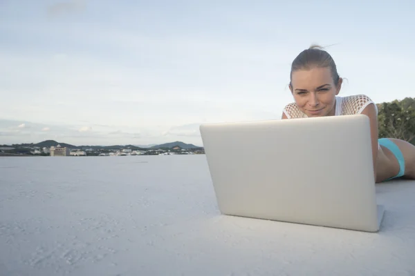 Outdoors portrait of pretty blonde woman in bikini with laptop computer lying on a rooftop over citylandscape and sky background. Freelance and technology