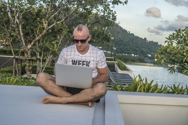 Outdoors portrait of handsome young man in sunglasses holding laptop computer on his knees while sitting on a rooftop over infinity swimming pool and sky background. Freelance and technology