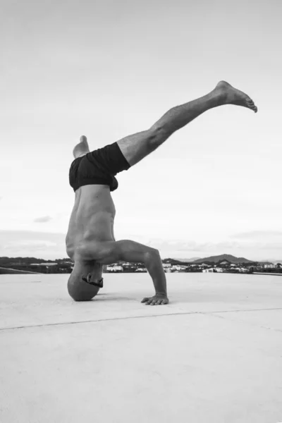 Black and white photo of handsome young man with naked torso and sunglasses doing brake dancing movements on a rooftop over sky background