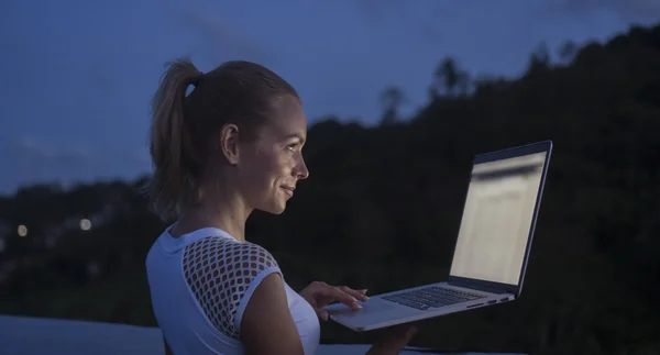 Outdoors portrait of pretty blonde happy woman with laptop computer in a rooftop infinity swimming pool over blurred mountain and sky background during evening. Freelance and technology