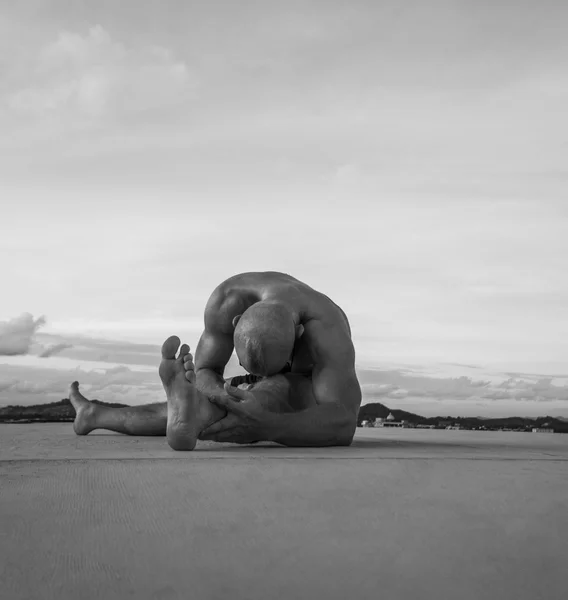 Black and white photo of handsome young man with naked torso doing stretching exercises on a rooftop over sky background