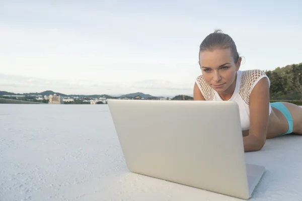 Outdoors portrait of pretty blonde woman in bikini with laptop computer lying on a rooftop over citylandscape and sky background. Freelance and technology