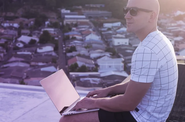 Outdoors portrait of handsome young man in sunglasses holding laptop computer on his knees while sitting on a rooftop over citylandscape. Freelance and technology