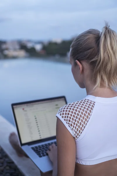 Back view of blonde woman in white top with laptop computer on her knees while sitting on a rooftop infinity swimming pool. Freelance and technology