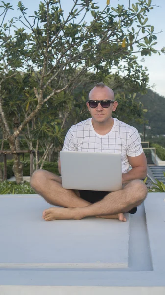 Outdoors portrait of handsome young man in sunglasses holding laptop computer on his knees while sitting on a rooftop over tree and and sky background. Freelance and technology
