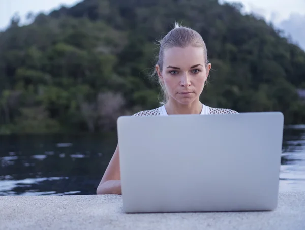 Outdoors portrait of pretty blonde woman in white top with laptop computer standing in a rooftop swimming pool over blurred green mountain and sky background. Freelance and technology