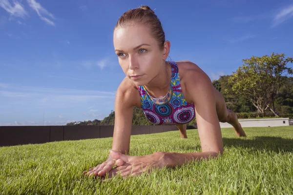 Bottom front view of pony tailed pretty blonde woman wearing colourful bikini doing plank position on a green grass rooftop over blue sky. Sport and healthy lifestyle concept