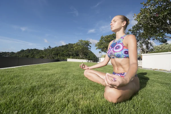 Bottom side view of pony tailed calm woman wearing colourful bikini meditating sitting on a green grass rooftop with closed eyes over blue sky. Sport and healthy lifestyle concept