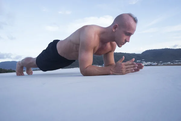 Bottom side view of handsome muscular man doing plank push up exercise on a rooftop over blue sky and green mountains tops. Sport and healthy lifestyle concept