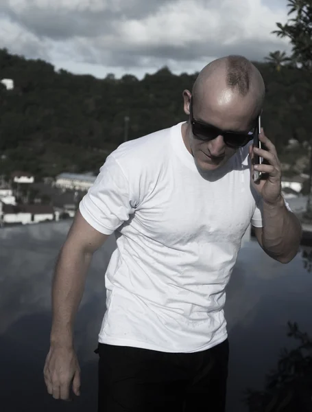 Outdoors portrait of handsome young man in sunglasses talking on the mobile phone while standing on a rooftop infinity swimming pool over over blue sky and city landscape. Freelance and technology