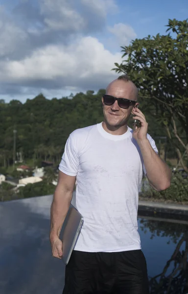 Outdoors portrait of cheerful young man in sunglasses talking on the mobile phone and holding laptop computer in his hand while standing on a rooftop infinity swimming pool over over blue sky and city landscape. Freelance and technology