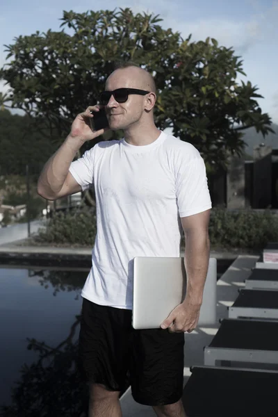 Outdoors portrait of handsome young man in sunglasses talking on the mobile phone and holding laptop computer in his hand while standing on a rooftop infinity swimming pool over over blue sky and city landscape. Freelance and technology