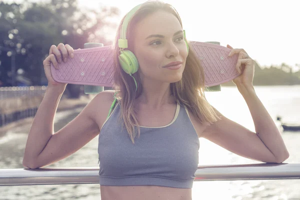 Young pretty blonde fitness female wearing bright green headphones listening music and holding pink skateboard while standing on the pier near the sea, enjoying early morning with beautiful sunrise