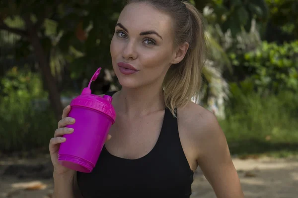 Close up of young healthy female wearing black exercise clothing holding pink shaker in her hand. Pretty happy blonde woman taking a break standing on on tropical beach during beautiful summer day