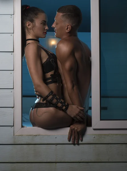 Young sexual lover couple of muscular handsome man and attractive brunette woman wearing black lingerie and swordbelts sitting and embracing in the  window during evening