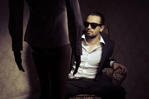 Young and elegant man sitting in chair wearing black suit, white shirt and sunglasses looking at woman standing in front of him isolated over vintage background. Back view silhouette of woman standing opposite handsome male in a retro inrerior