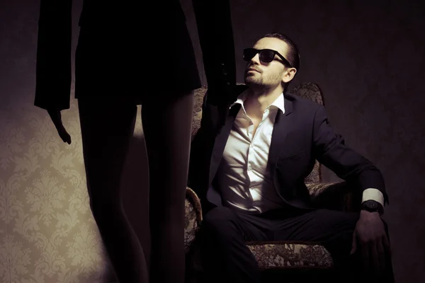 Young and elegant man sitting in chair wearing black suit, white shirt and sunglasses looking at woman standing in front of him isolated over vintage background. Back view silhouette of woman standing opposite handsome male in a retro inrerior