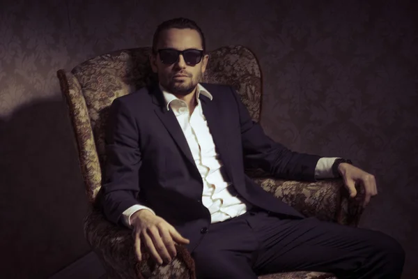 Young handsome and elegant man sitting in chair wearing black suit and sunglasses isolated over vintage background