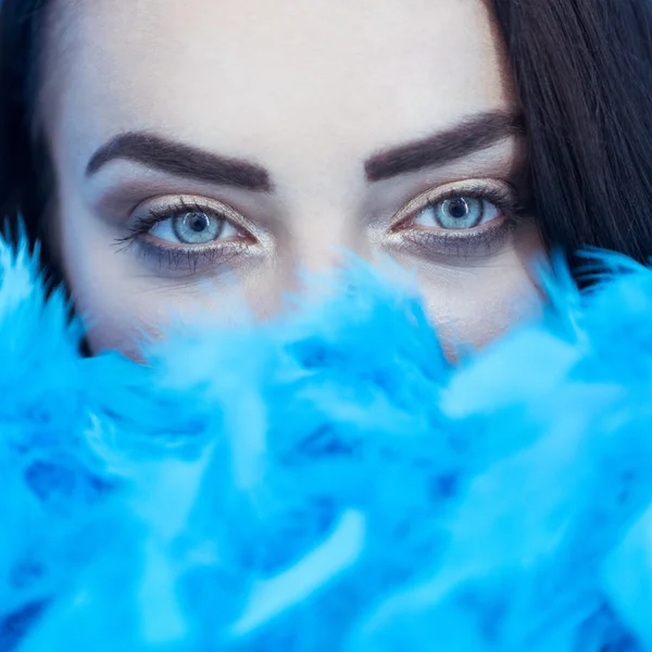 Closeup portrait of beautiful seductive brunette woman smiling and looking into the camera while playing with blue feather boa over blue wall background