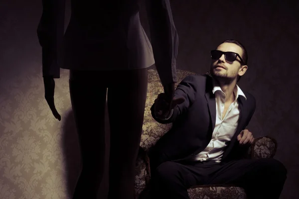 Young and elegant man sitting in chair wearing black suit, white shirt and sunglasses looking at woman standing in front of him isolated over vintage background