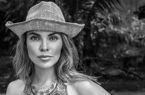 Portrait of beautiful young woman with straw hat on a sunny day looking into camera over palm trees background. Black and white photo - eyes closeup of pretty girl with straw hat pattern shadow on her face