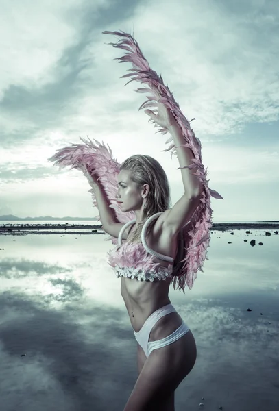 Romantic young beauty as an angel on the beach. Side view of blonde pretty female woman wearing pink wings, feather bra and bikini bottom standing in the sea over cloudy sky background