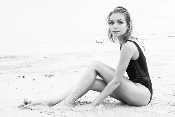 Black and white photo of elegant blonde pretty woman with wet look wearing black swimsuit sitting on the sandy beach near the sea over sky background