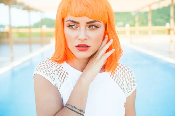 Closeup portrait of sexy beautiful woman in modern futuristic style posing by the rooftop pool during sunny summer day. Creative look of tattooed woman wearing white bikini and orange wig