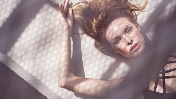 Closeup portrait of beautiful blonde glamour woman wearing black swimwear lying down on a rooftop in a sunny summer day with shadow net reflection from metal sunshade on her face