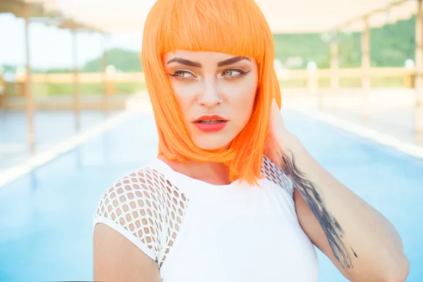 Closeup portrait of sexy beautiful woman in modern futuristic style posing by the rooftop pool during sunny summer day. Creative look of tattooed woman wearing white bikini and orange wig